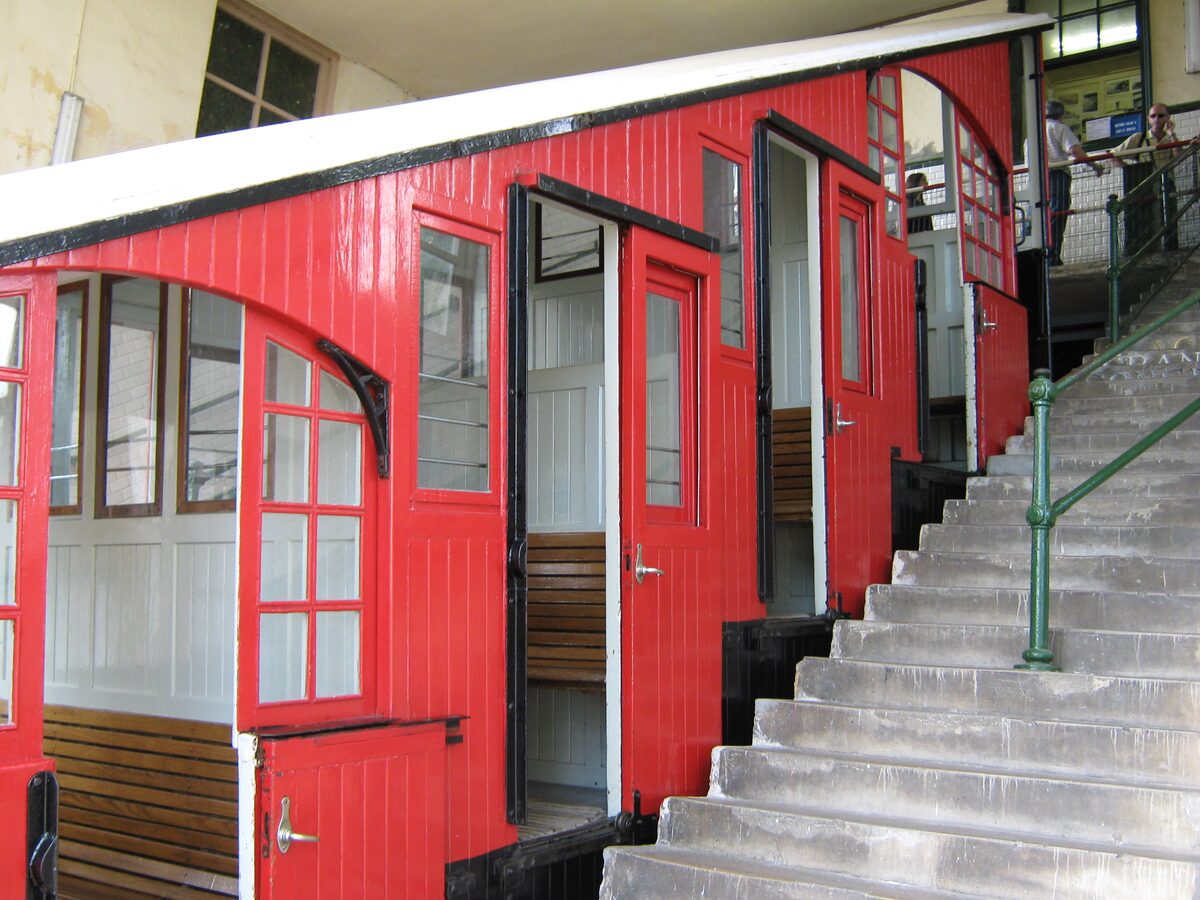 A red wooden funicular car at the bottom station of the Igueldo funicular, with a stone staircase running beside it