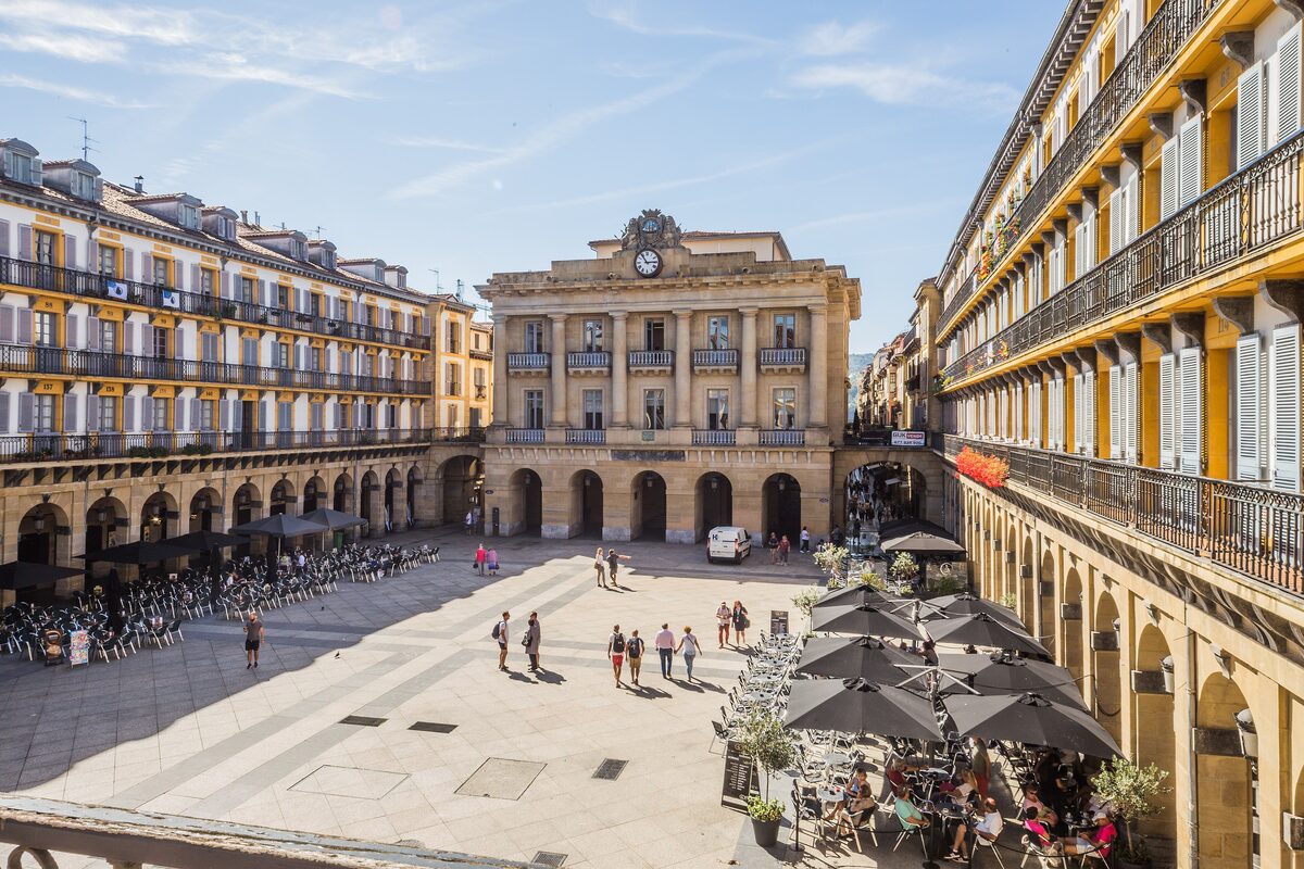 Plaza de la Constitución seen from above: the arcaded square with cafe tables, the old town hall building at the far end and rows of balconies on the yellow and white facades