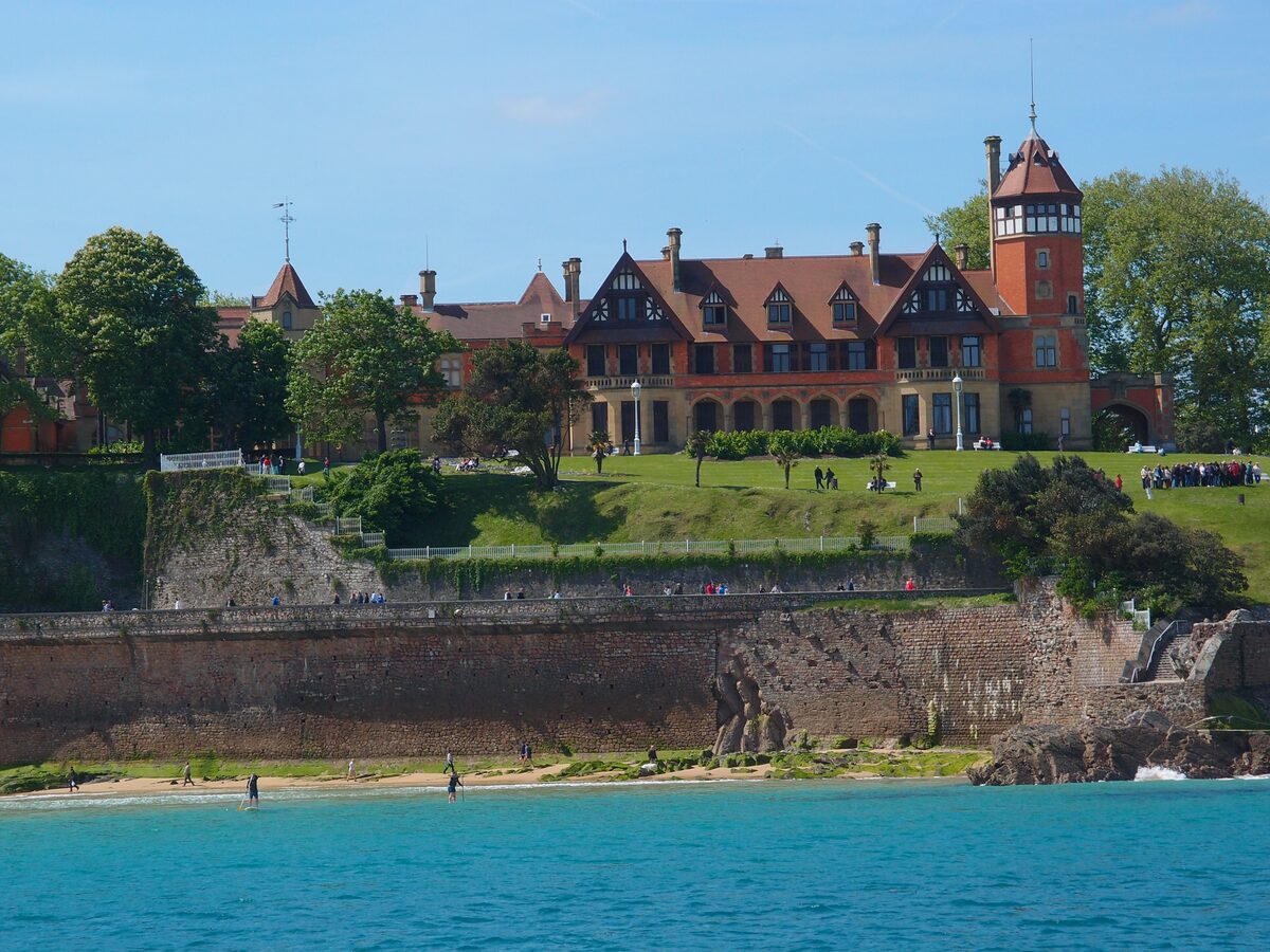 Miramar Palace, a red-brick and sandstone villa with a tower, seen above a sea wall with the turquoise water below