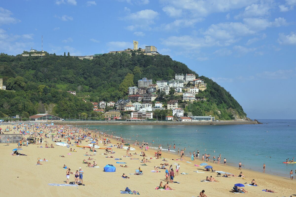 Ondarreta beach in the afternoon, with sand and sunbathers, the water of the bay in front and the wooded hill of Igueldo behind the town