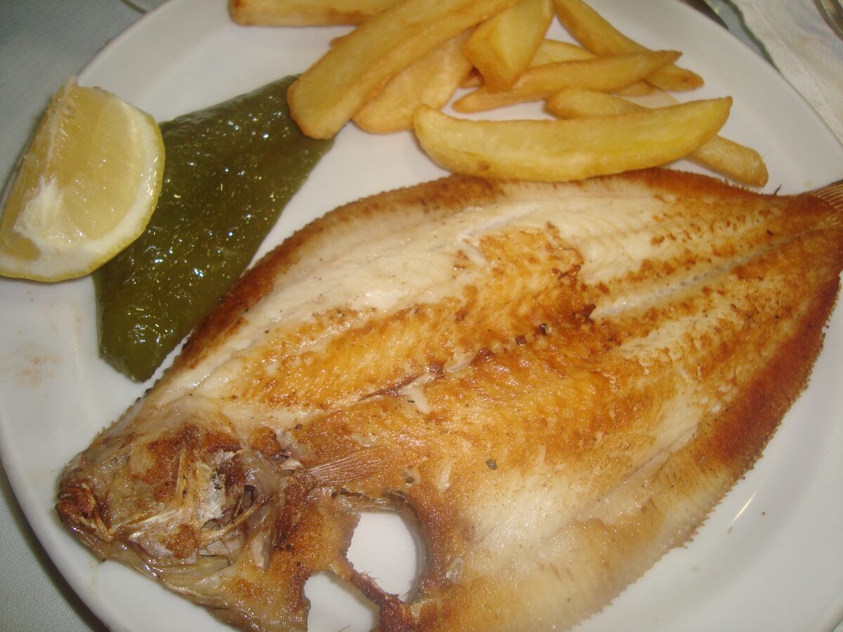 A grilled flatfish on a white plate, with a lemon wedge and fried potatoes