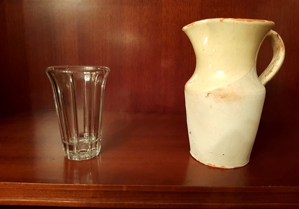 A glass and a cream ceramic pitcher of the kind used at txakoli wineries, on a wooden shelf