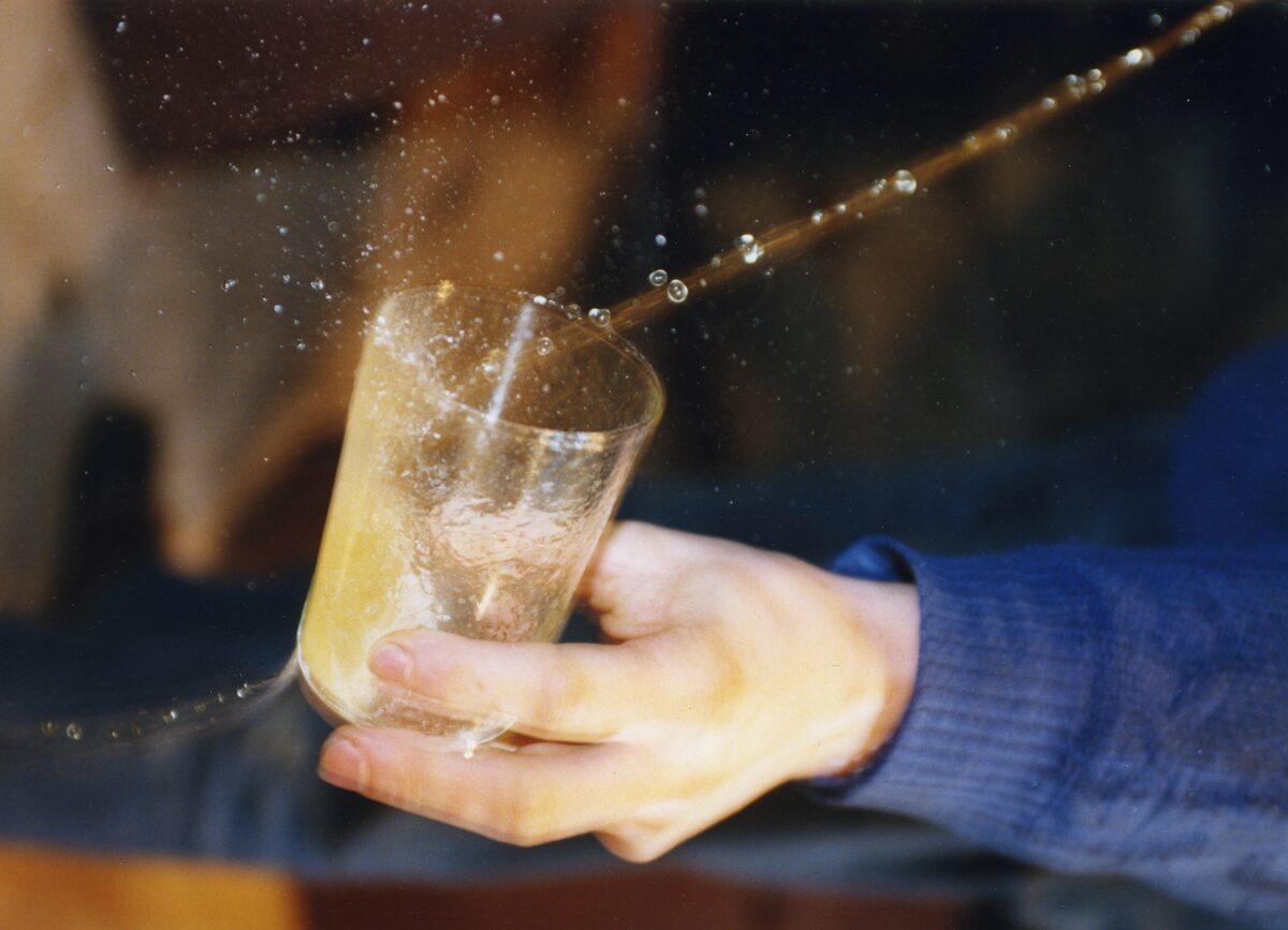 A hand holds a glass under a thin jet of cider from a barrel, the liquid splashing as it fills