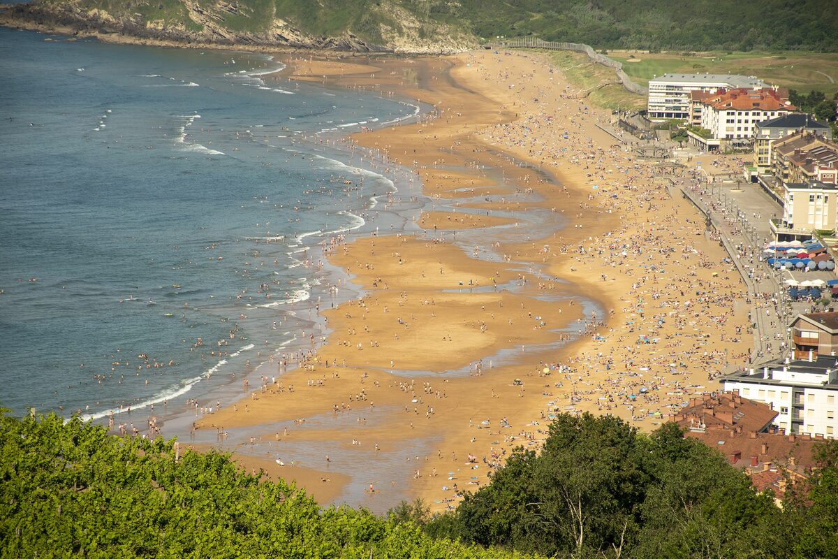 Zarautz beach from a hillside viewpoint: a long strip of sand curving along the bay, with the town's buildings at the right edge