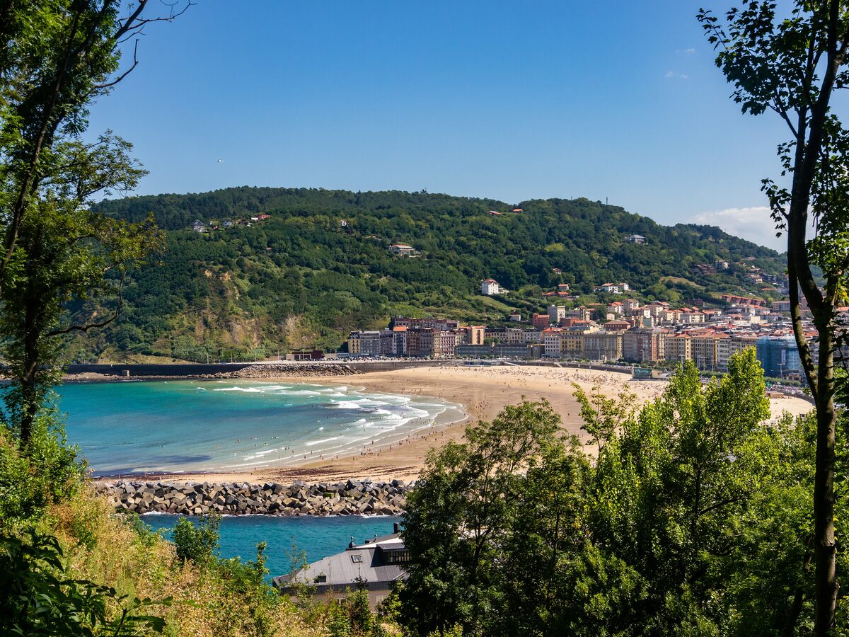 Zurriola beach seen through trees: surf on the sand, a stone breakwater, the buildings of Gros and wooded Monte Ulia behind