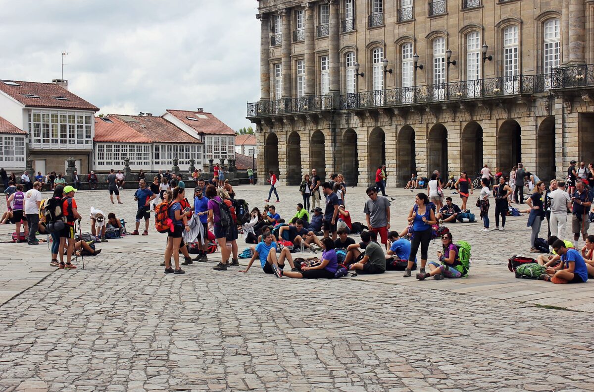 Pilgrims with backpacks resting on the paving of the Praza do Obradoiro, with the arcades of the Pazo de Raxoi behind