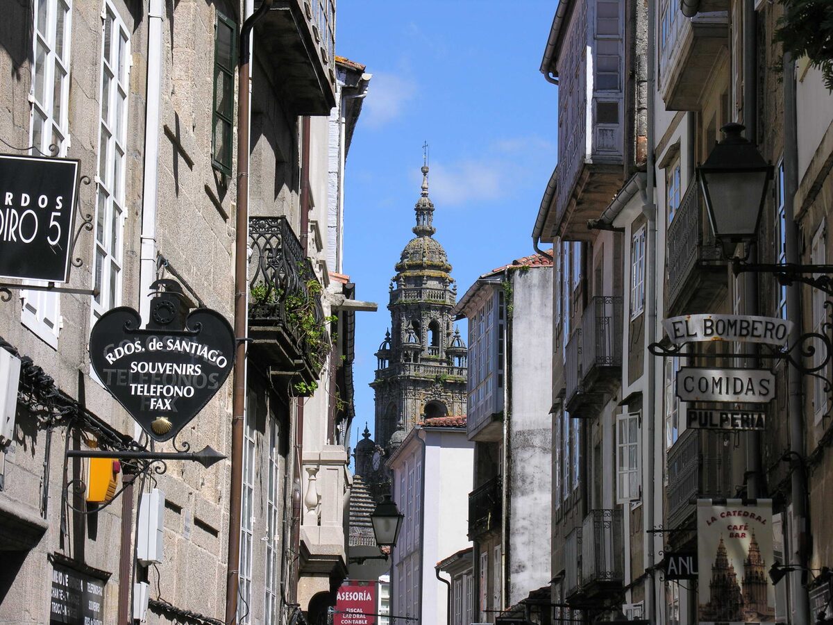 A narrow old-town lane of Rúa do Franco with hanging bar signs, one reading Comidas, and a cathedral tower against blue sky