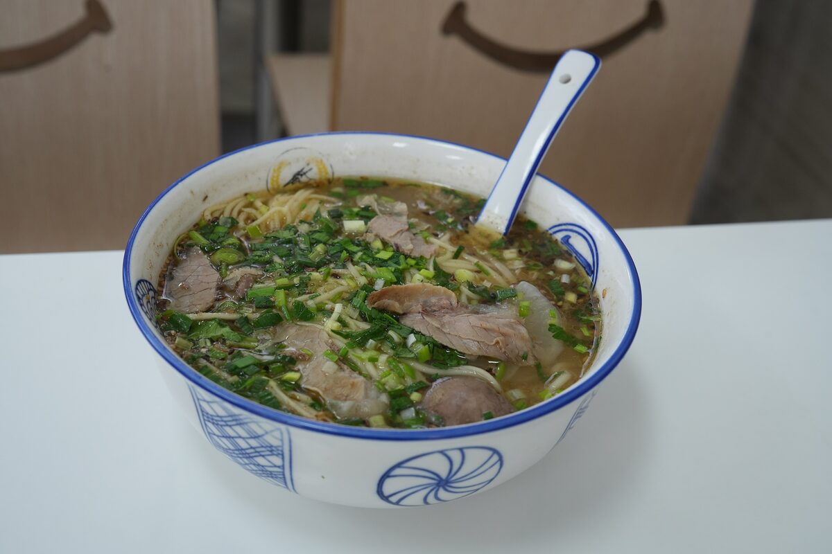 A blue-and-white bowl of beef noodle soup with sliced beef, scallions and a spoon