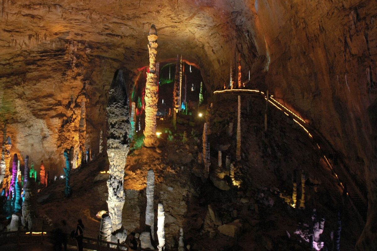 A lit cavern in Huanglong Cave with tall stalagmites in colored light and a railed walkway along the wall