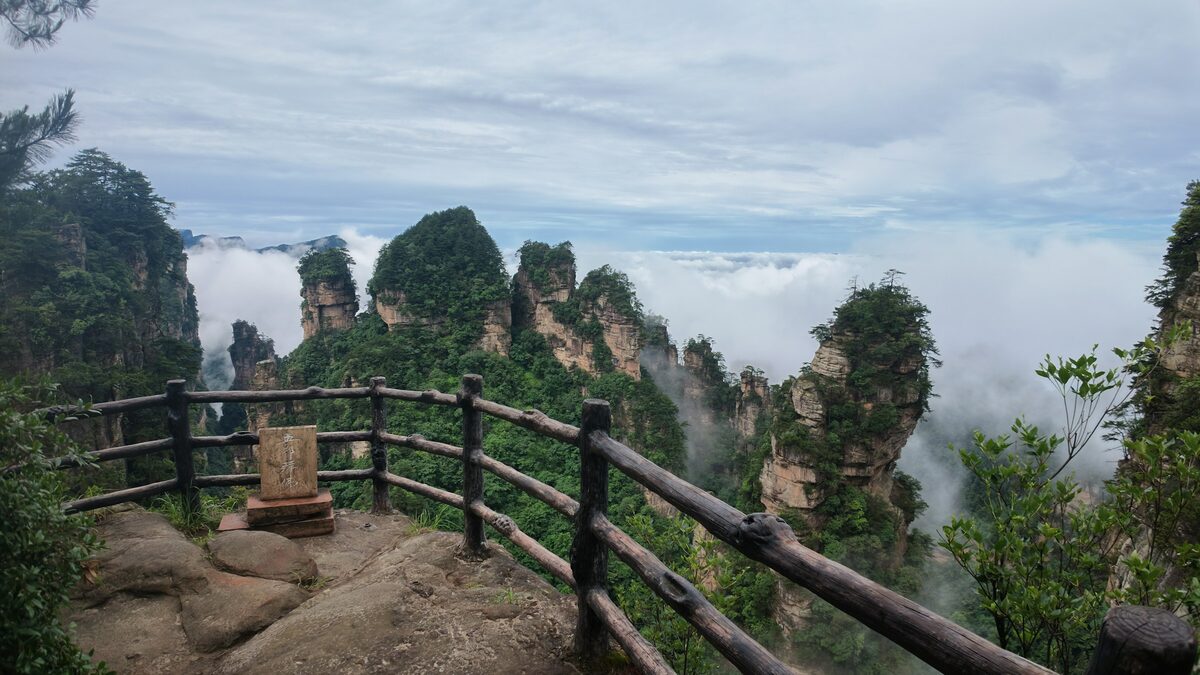 A wooden railing at a rocky lookout over cloud-filled valleys with pillar-shaped peaks in the distance