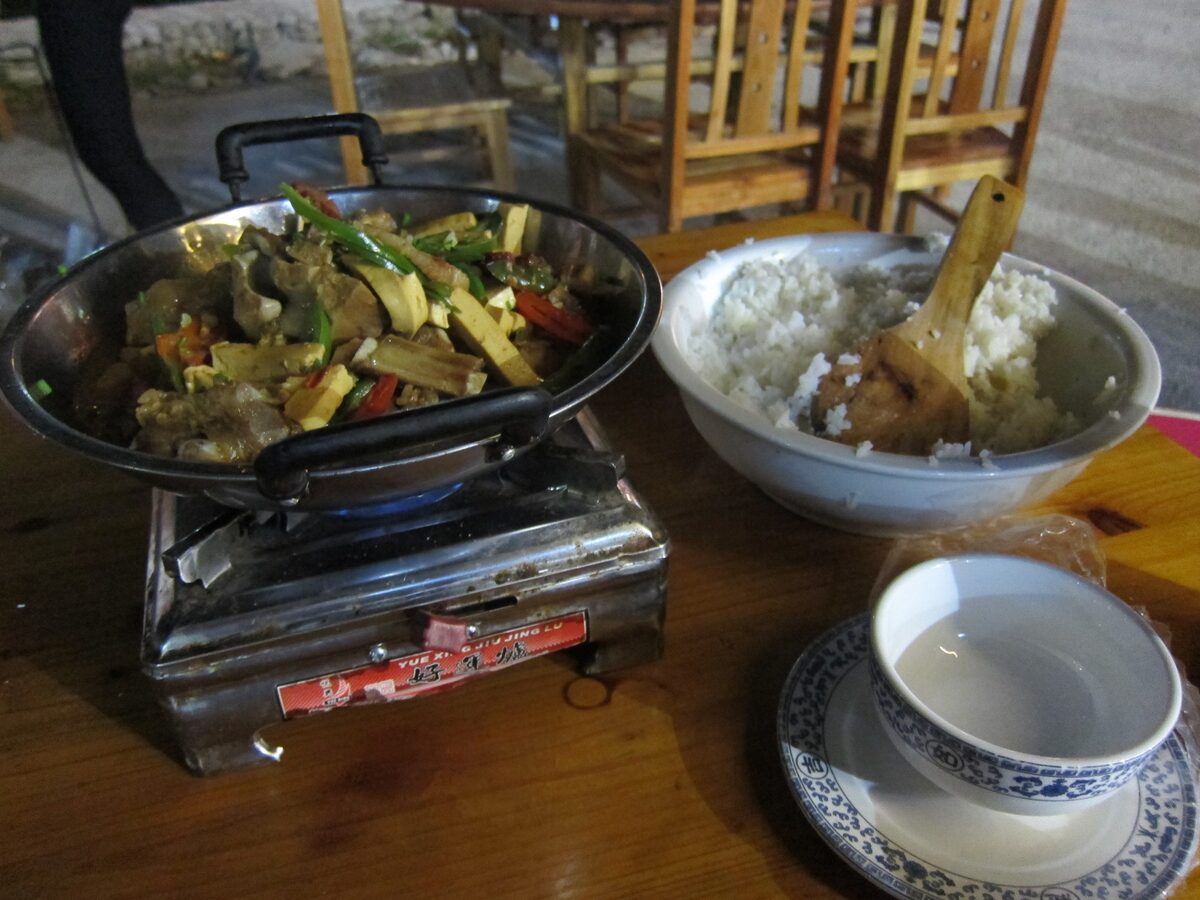 A wok of meat and vegetables simmering on a tabletop burner beside a bowl of rice and a small cup