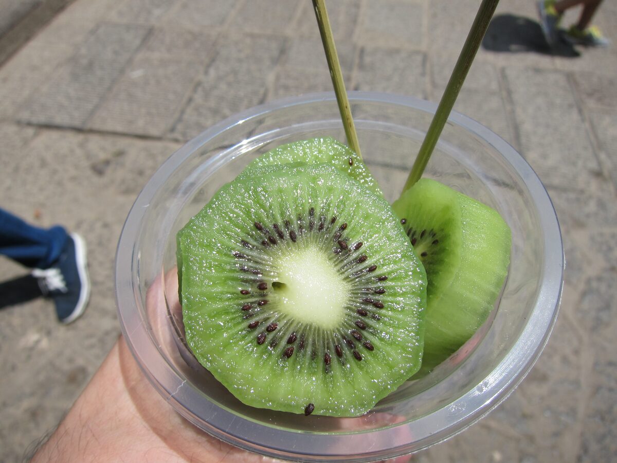 A hand holding a clear cup of sliced kiwi with two bamboo skewers, a fruit snack from a street stall in the Wulingyuan area
