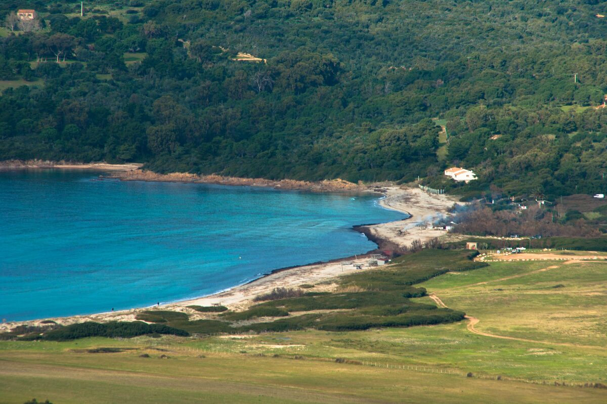 Capo di Feno from above: a bay of turquoise water with a pale sand strip, wooded hills and green fields behind
