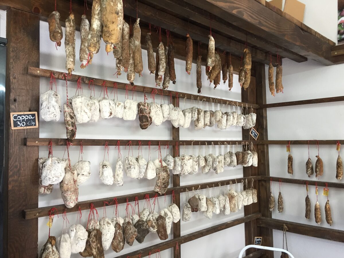 Dry-cured sausages and cured meats hanging on wooden racks in a Corsican charcuterie shop, with price tags for coppa and lonzu