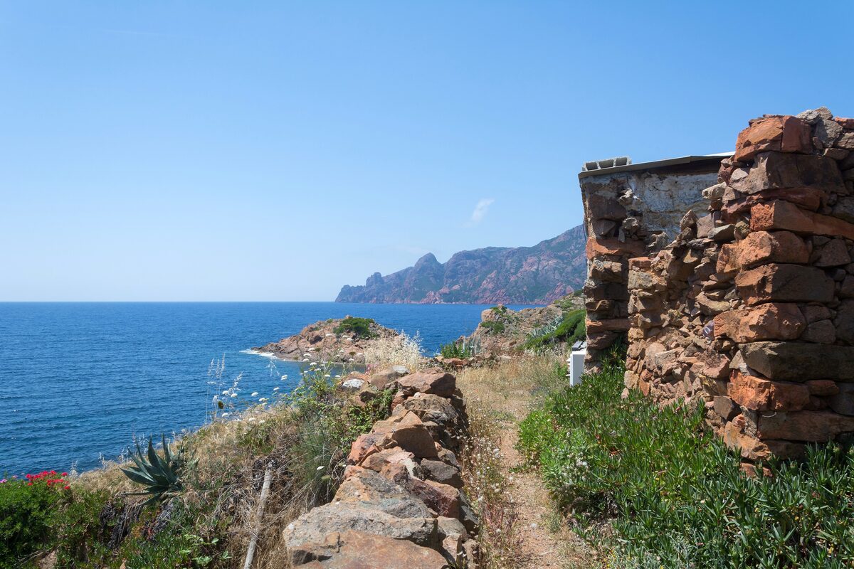 Stone ruins beside the coastal path at Girolata, with the sea and the red cliffs of Scandola beyond