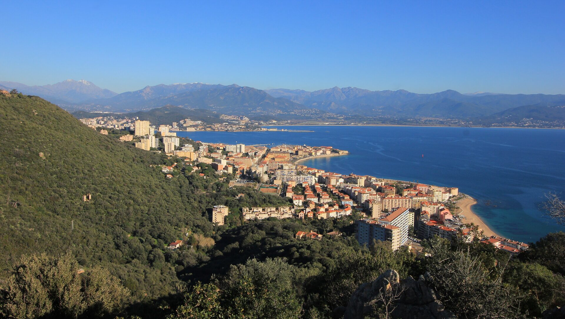 Ajaccio and its gulf from the hills to the west on a clear day: the old town on its point, a sandy beach, deep blue water and the mountains of the interior behind