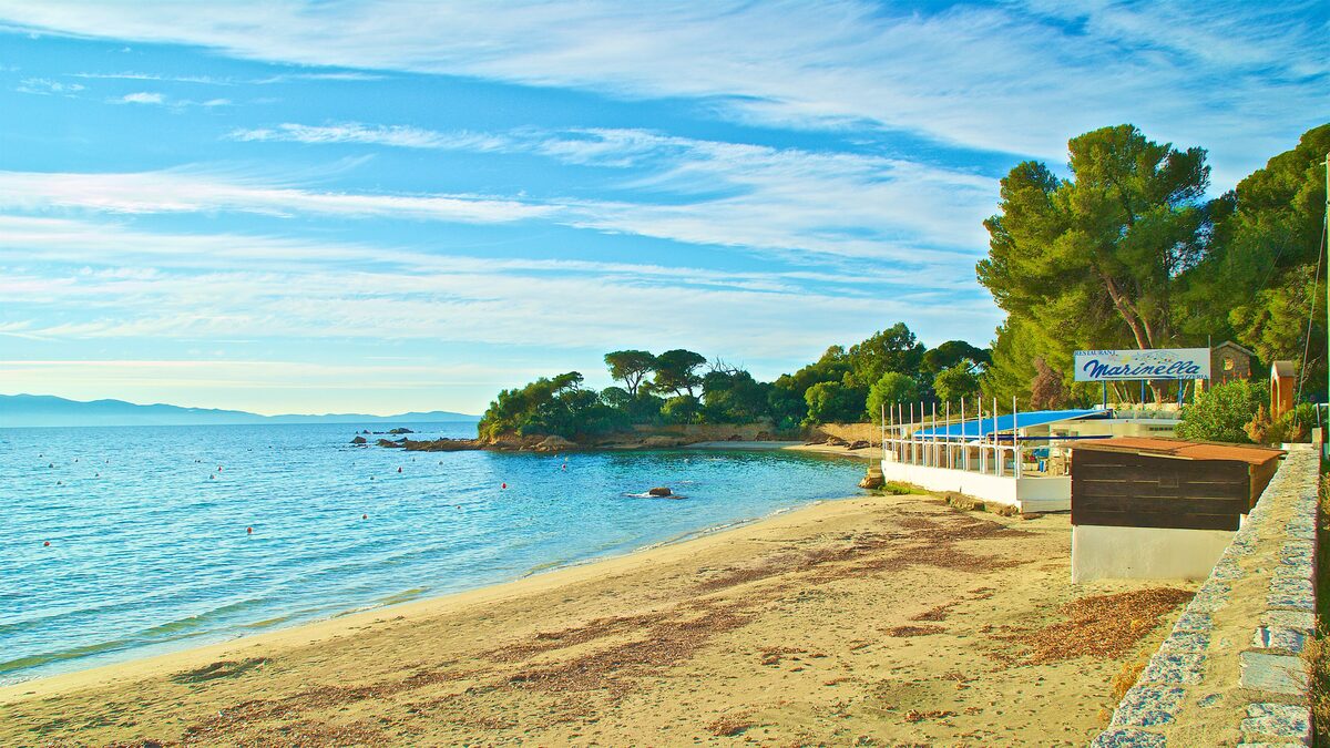 Marinella beach on a clear morning: a strip of sand beside calm turquoise water, with pines and a beach restaurant on the right
