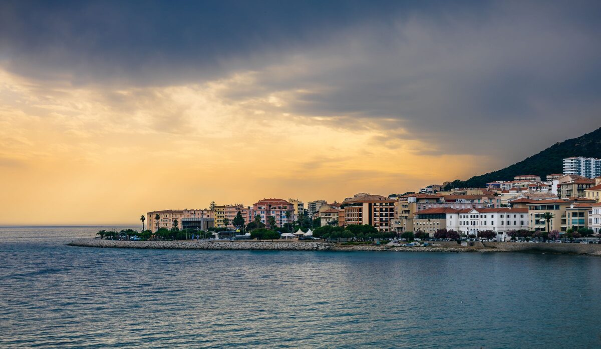 The Ajaccio waterfront and its breakwater seen across the water at dusk, with golden light under a heavy sky and apartment blocks along the shore