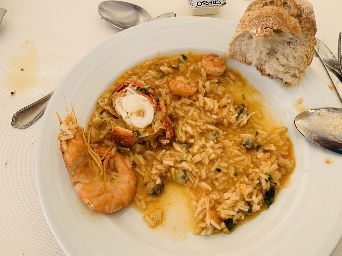 A plate of soupy seafood rice with a prawn, lobster tail and clams, and a piece of bread