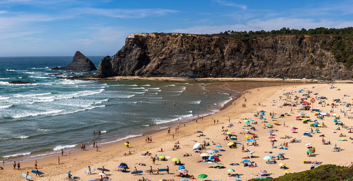 Odeceixe beach on a sunny day, with sand, surf and a cliff at the end of the bay