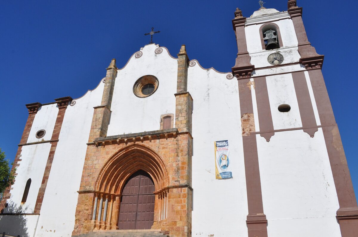 The white front and bell tower of the Sé de Silves with its red sandstone Gothic doorway under a blue sky