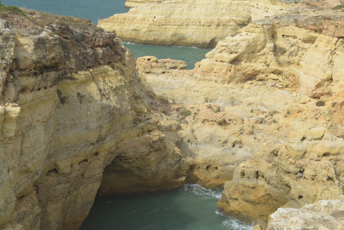 Golden sandstone cliffs with a sea cave above green water, seen from the Seven Hanging Valleys trail near Praia do Carvalho