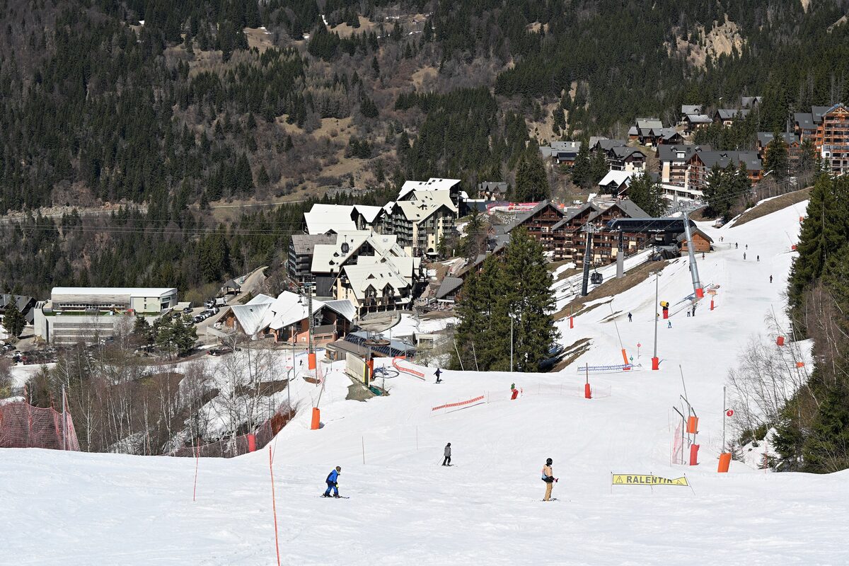The Oz 3300 pistes in snow with skiers, the village buildings at the foot of the slope and the mountains behind