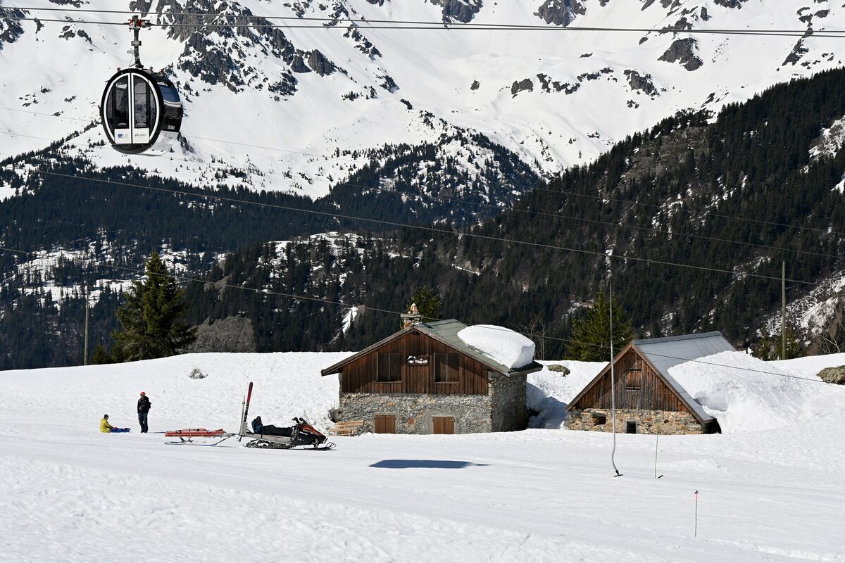 A gondola cabin above two stone-and-wood mountain chalets in the snow near Montfrais, with a snowmobile and a sled in front