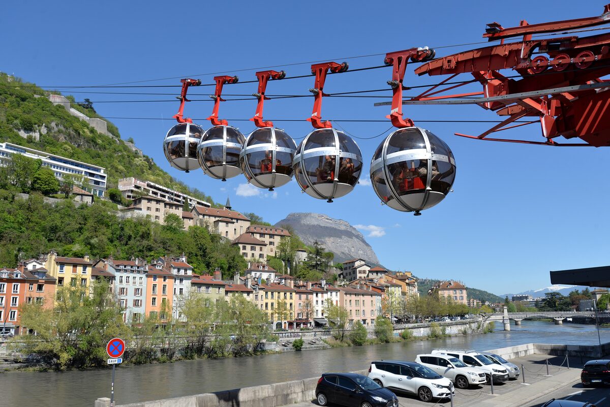 A row of Bastille cable car bubbles over the Isère river, with the houses of Grenoble and green hillsides in spring light