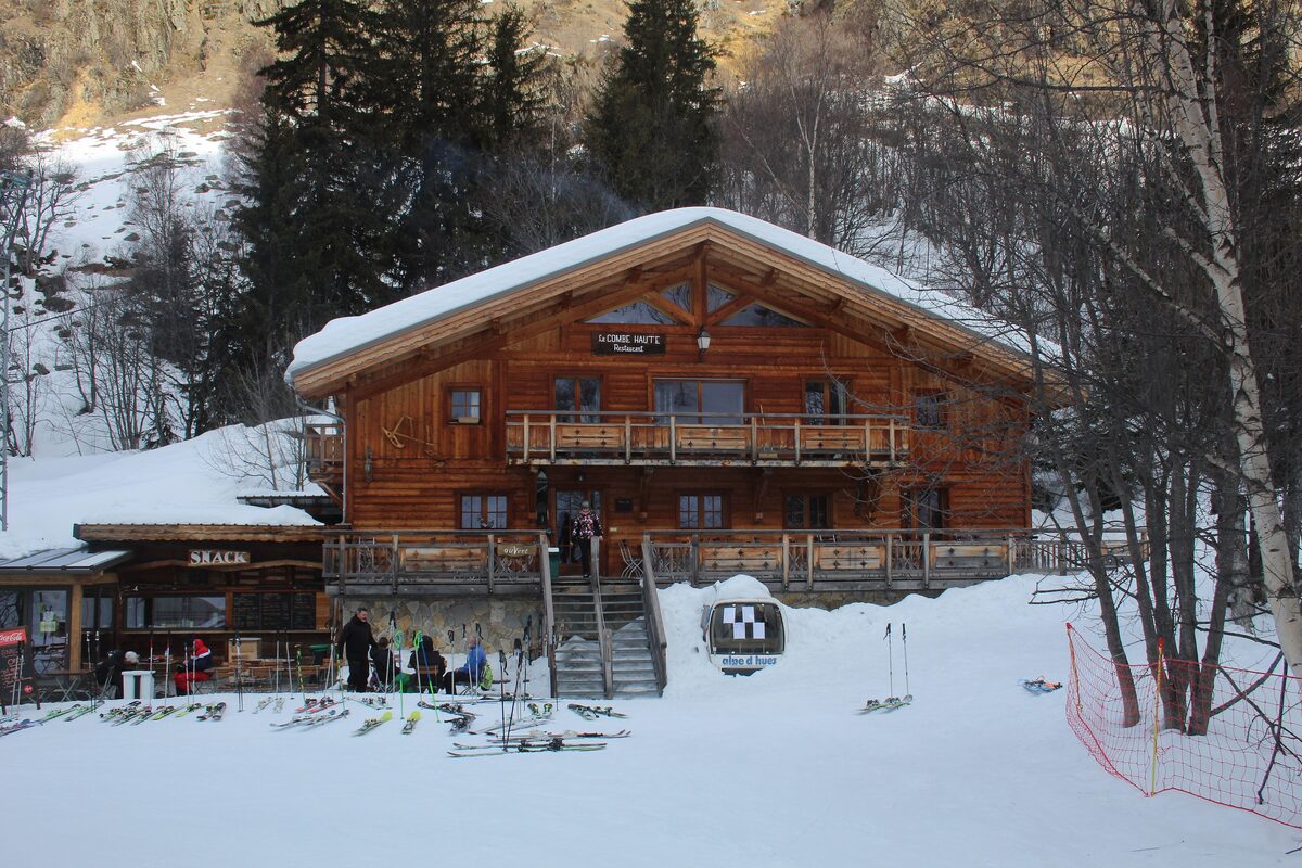 The wooden Auberge de la Combe Haute in the snow, with skiers and skis outside the terrace