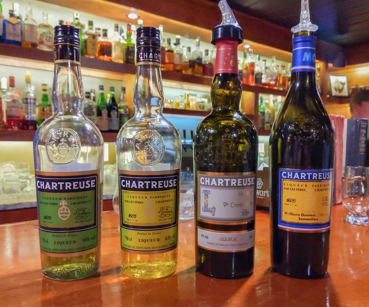 Four bottles of Chartreuse liqueur with different labels, standing on a bar with shelves of bottles behind