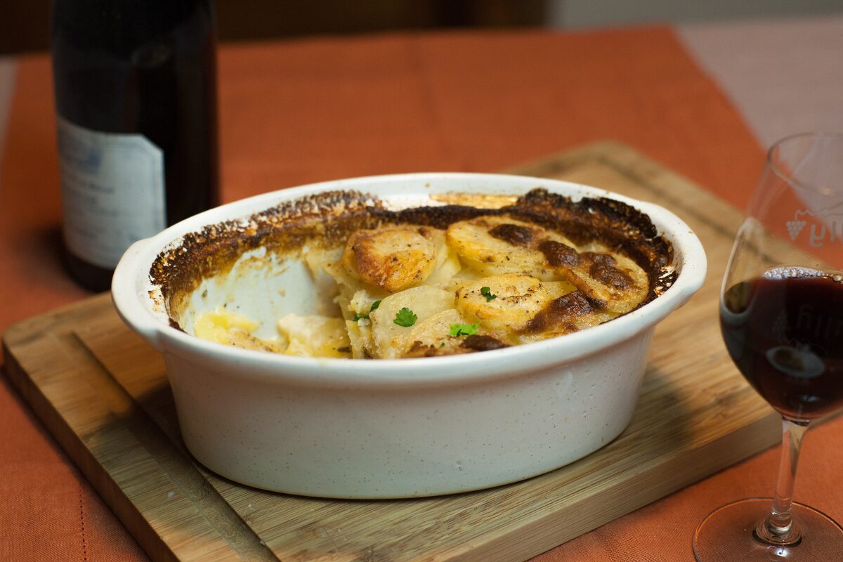 A gratin dauphinois in a white oval dish, golden on top, with a glass of red wine beside it