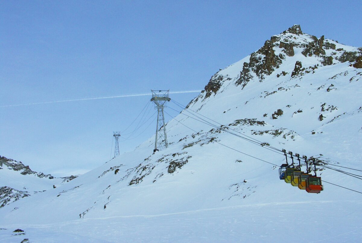 Colourful cabins of the La Grave cable car running above a snowy slope, with a pylon and a rocky summit behind