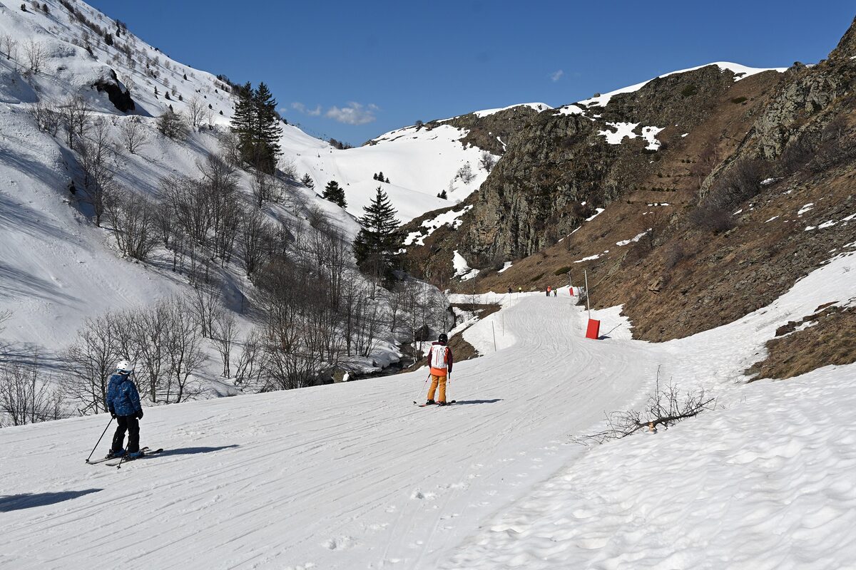 Two skiers on the wide Sarenne piste in a narrow valley, with bare trees and a stream on the left