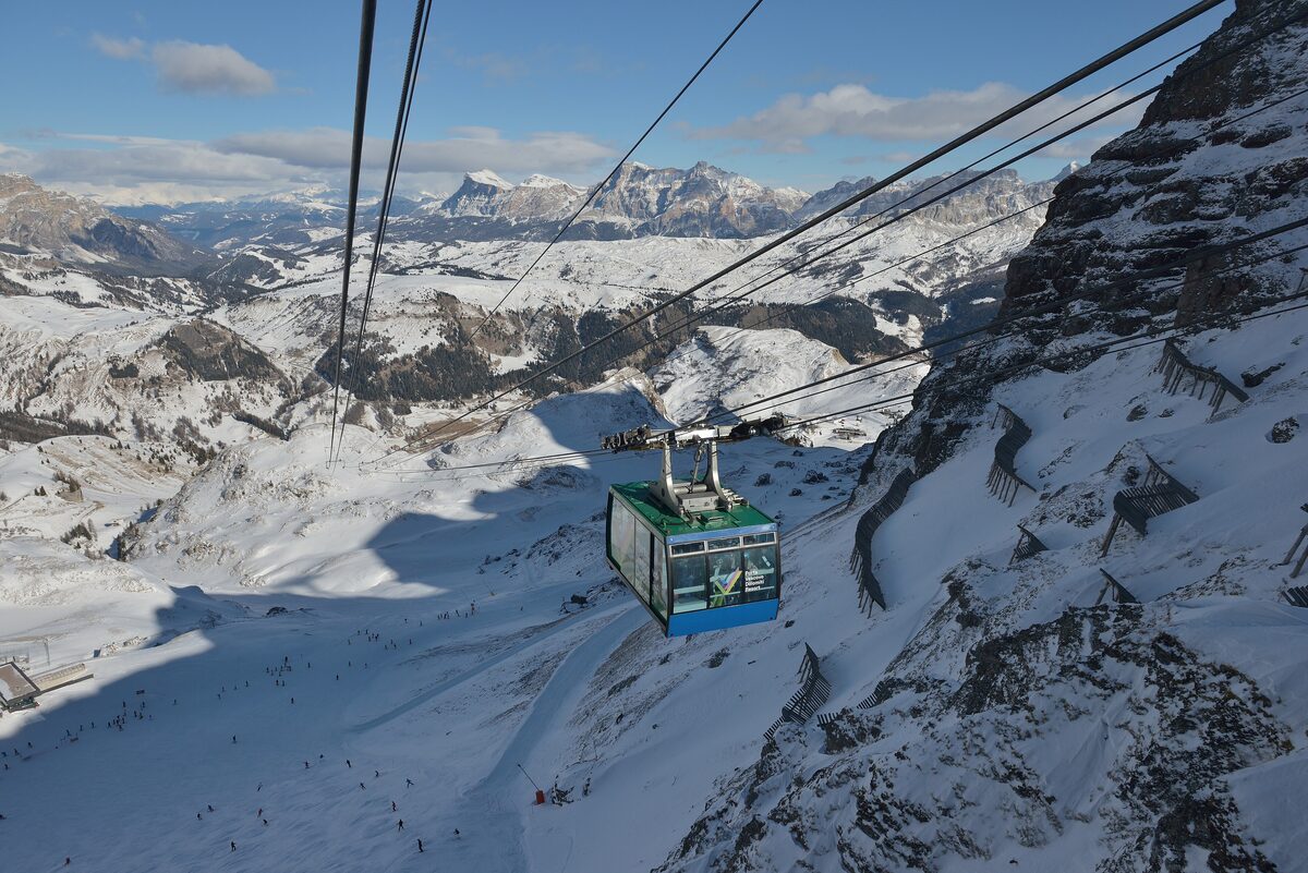 A cabin of the Porta Vescovo cable car crossing snowy slopes, with distant snow-capped peaks behind