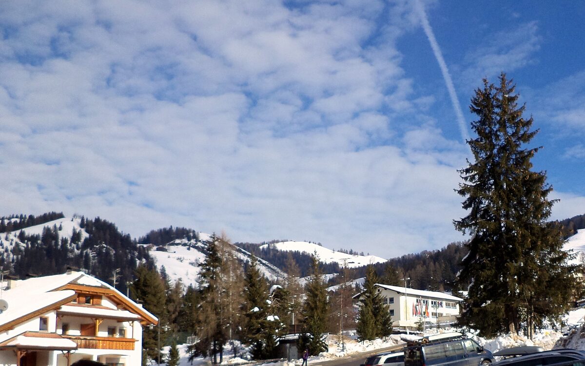 A snow-roofed chalet hotel and larches at Pedraces in Badia, with snowy hills behind under a sky of thin cloud