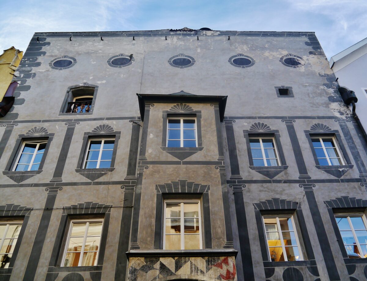 A grey house front with painted dark bands, a bay window and round windows in the Brunico old town