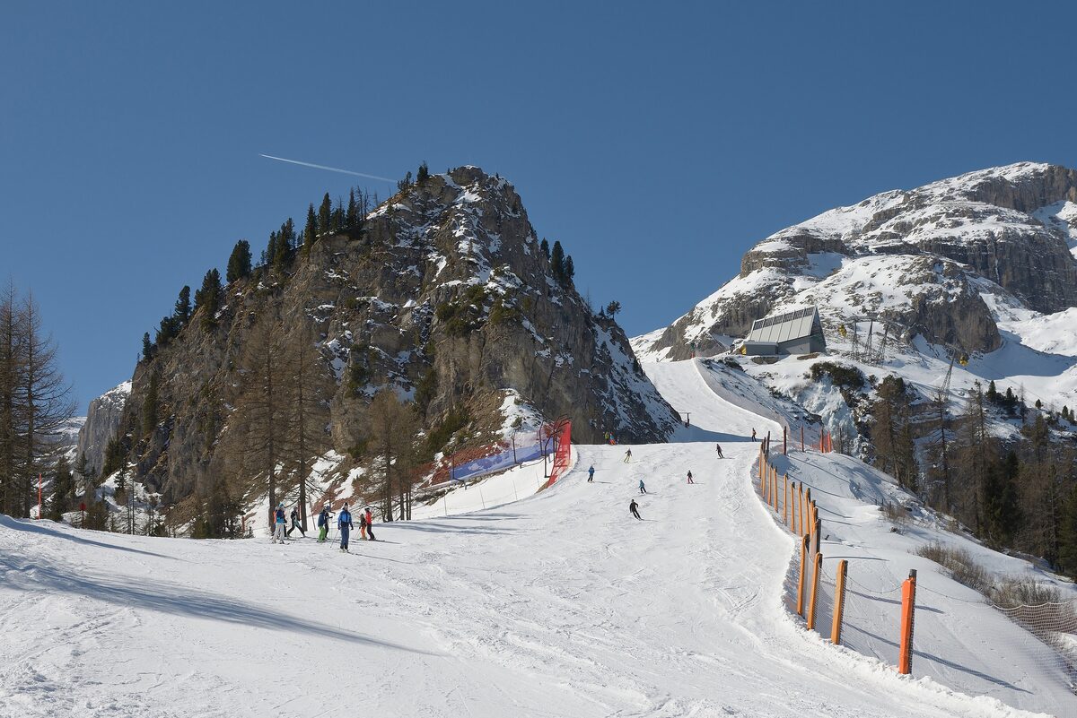 Skiers on a wide snowy piste below a rock tower, with larches along the slope and a lift station on the ridge to the right