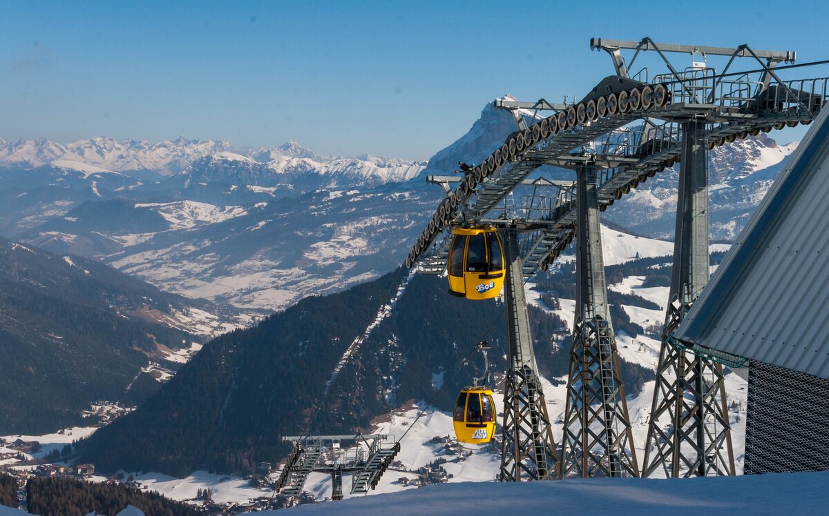 Two yellow Alta Badia gondola cabins, one marked Boè, passing a lift pylon above a snowy valley near Corvara on a clear day