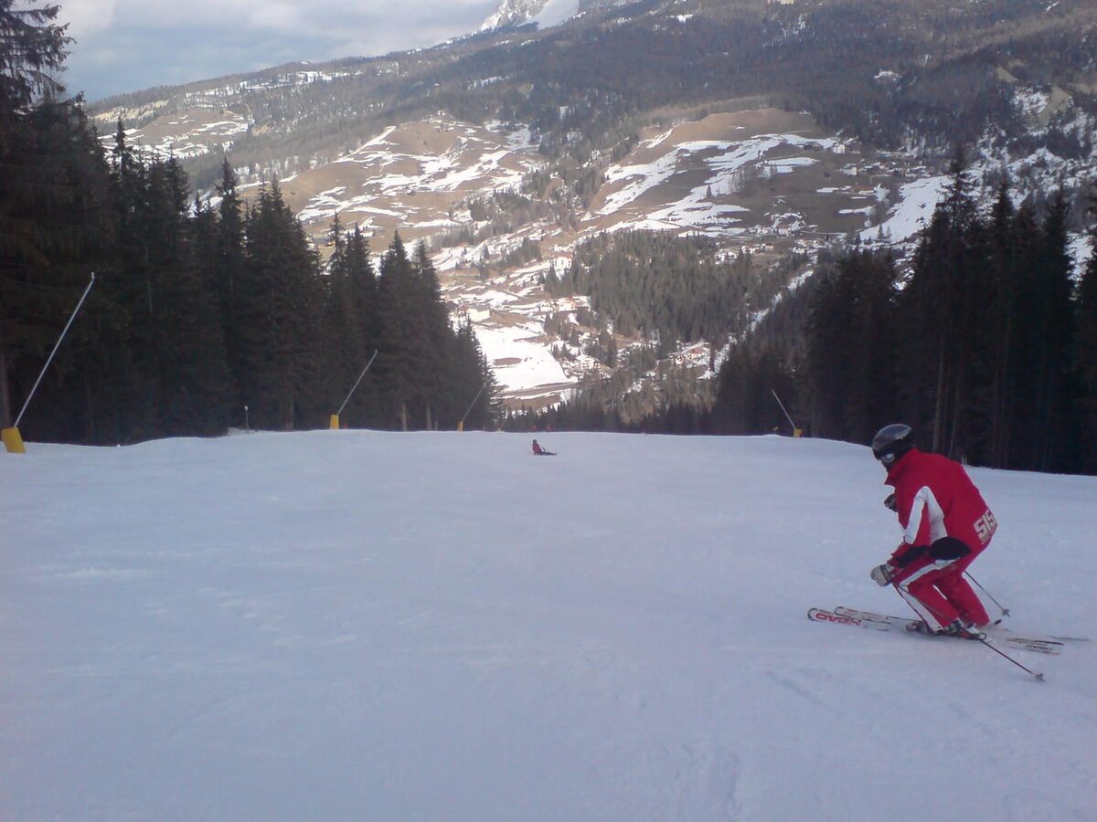A skier in a red suit crouching down a wide snowy piste above the forest, the Gran Risa seen from La Villa