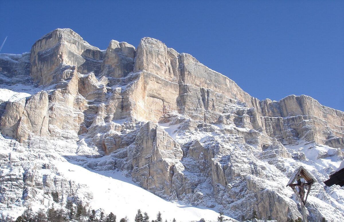 The pale rock walls of the Sas dla Crusc under a blue sky, snow on the ledges, with a wayside crucifix at the lower right (the gondolas are not in frame)
