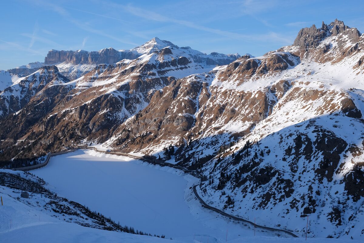 A frozen mountain reservoir under snow, with a high flat-topped rock massif beyond, a dam road curving along its shore