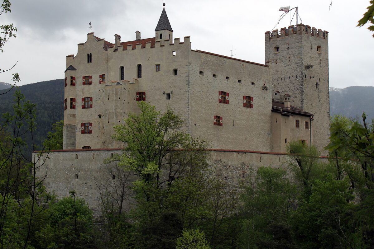 Bruneck Castle in spring, a pale fortress with crenellated towers and red shutters rising above green trees