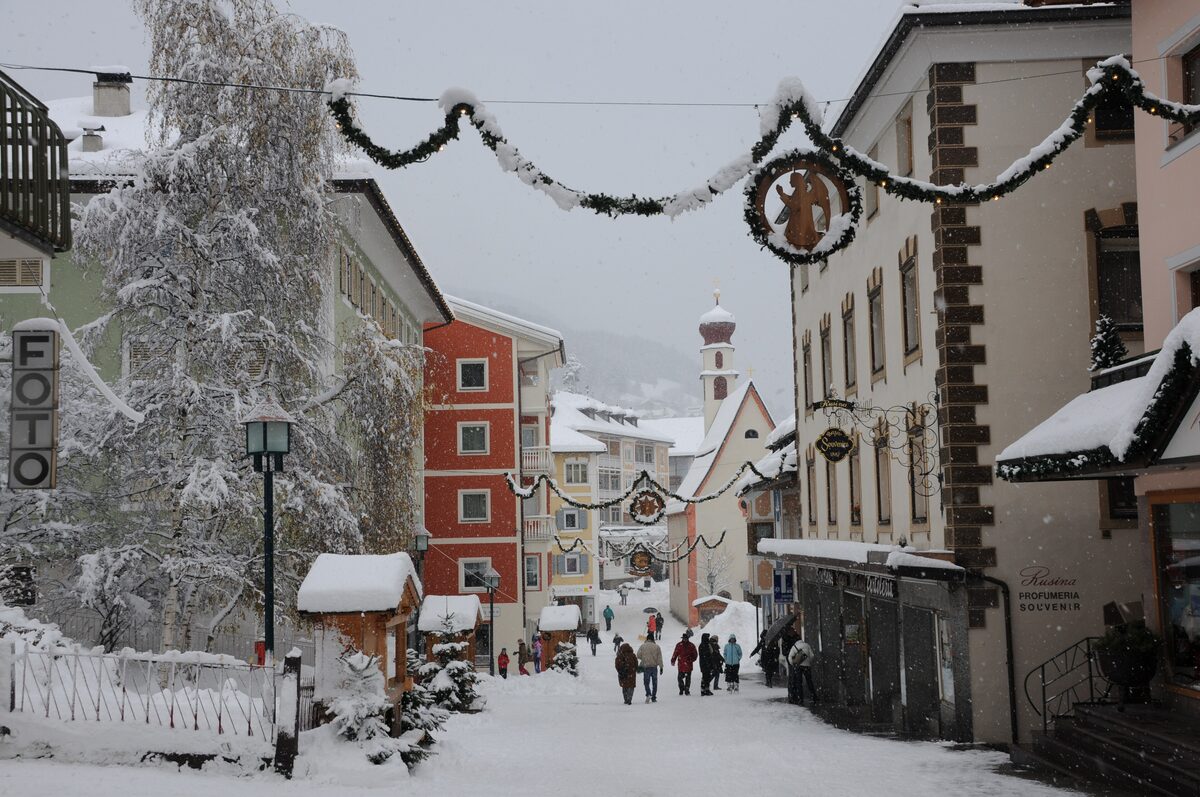 A snowy street in Ortisei with Christmas garlands on the old houses and people walking past the church