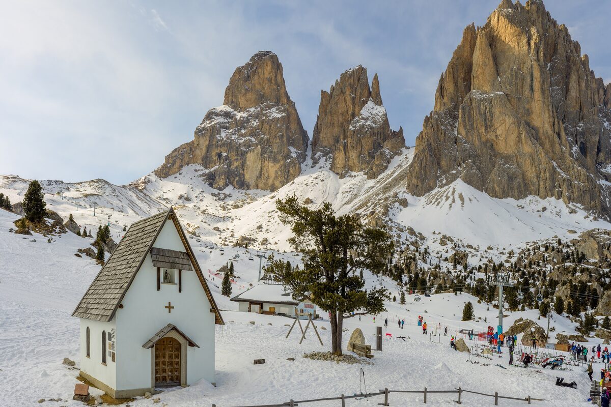 The white Passo Sella chapel in the snow with skiers by the lift and the Langkofel towers behind