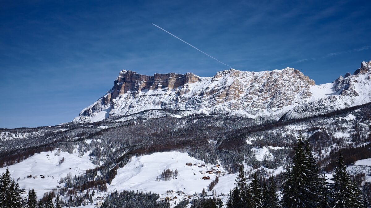 The Sas dla Crusc rock wall above dark forest and a snowy slope with a few chalets, on a clear winter day