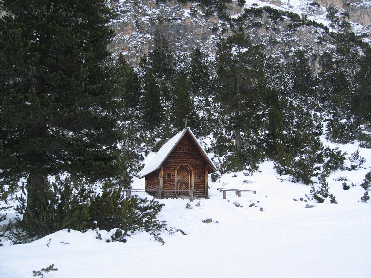 A small log memorial chapel with snow on its roof among pines, near the Scotoni hut (the hut itself is not in frame)