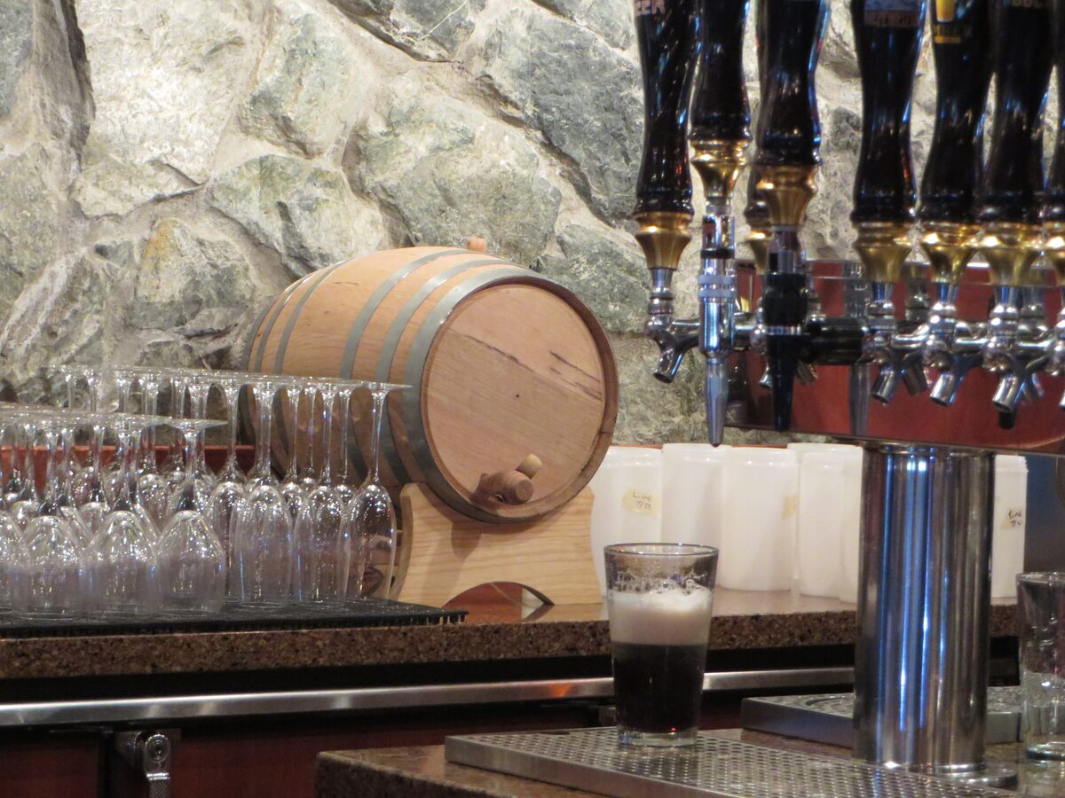 A pint of dark beer with a foam head beside a small oak cask, beer taps and rows of glasses at the Glacier BrewHouse