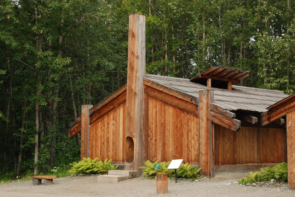 A replica Alaska Native house with carved posts and a timber roof among birch and spruce trees at the Heritage Center