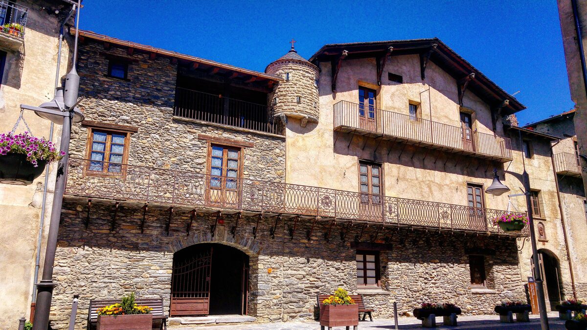 The stone and plaster facade of Casa d'Areny-Plandolit in Ordino, with a wrought-iron balcony, a small round corner tower and flower boxes on the square